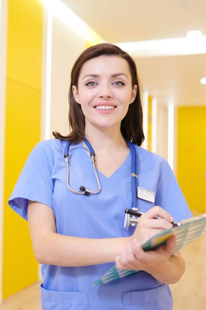 Waist up portrait of smiling nurse looking at camera while posing in clinic holding clipboard